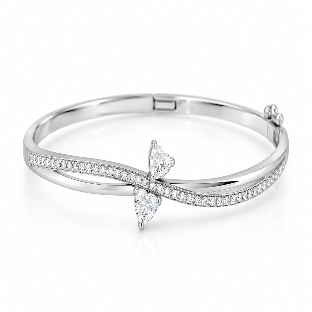 A silver-toned hinged bangle bracelet featuring a bow-shaped setting with two clear heart-cut gemstones and a row of small pavé-set stones along the curved upper band, shown on a white background.