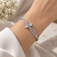 A silver-toned hinged bangle bracelet featuring a bow-shaped setting with two clear heart-cut gemstones and a row of small pavé-set stones along the curved upper band, shown on a white background.