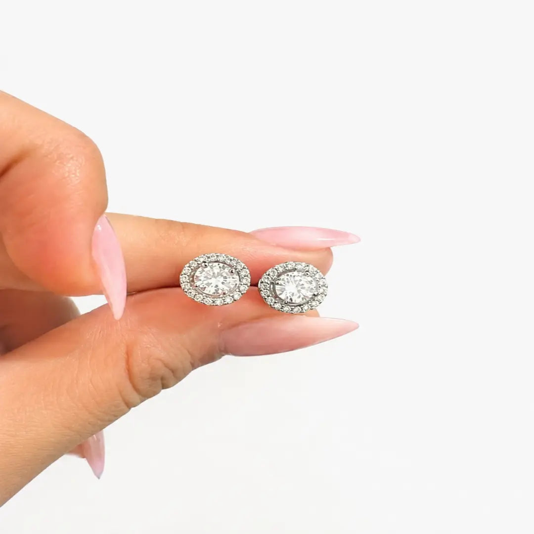 A close-up of a hand with pink manicured nails holding a pair of round halo stud earrings with clear gemstones on a plain white background.