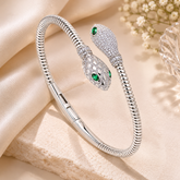 A silver snake-head bangle bracelet with pavé-set stones and green emerald eyes, displayed on a beige fabric surface with a small glass object in the background.