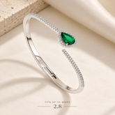 A polished silver-toned open cuff bracelet with a teardrop-shaped green gemstone surrounded by small clear stones, displayed on a soft cream fabric background with a textured block and text reading 'FITS UP TO WRIST 2.8'.
