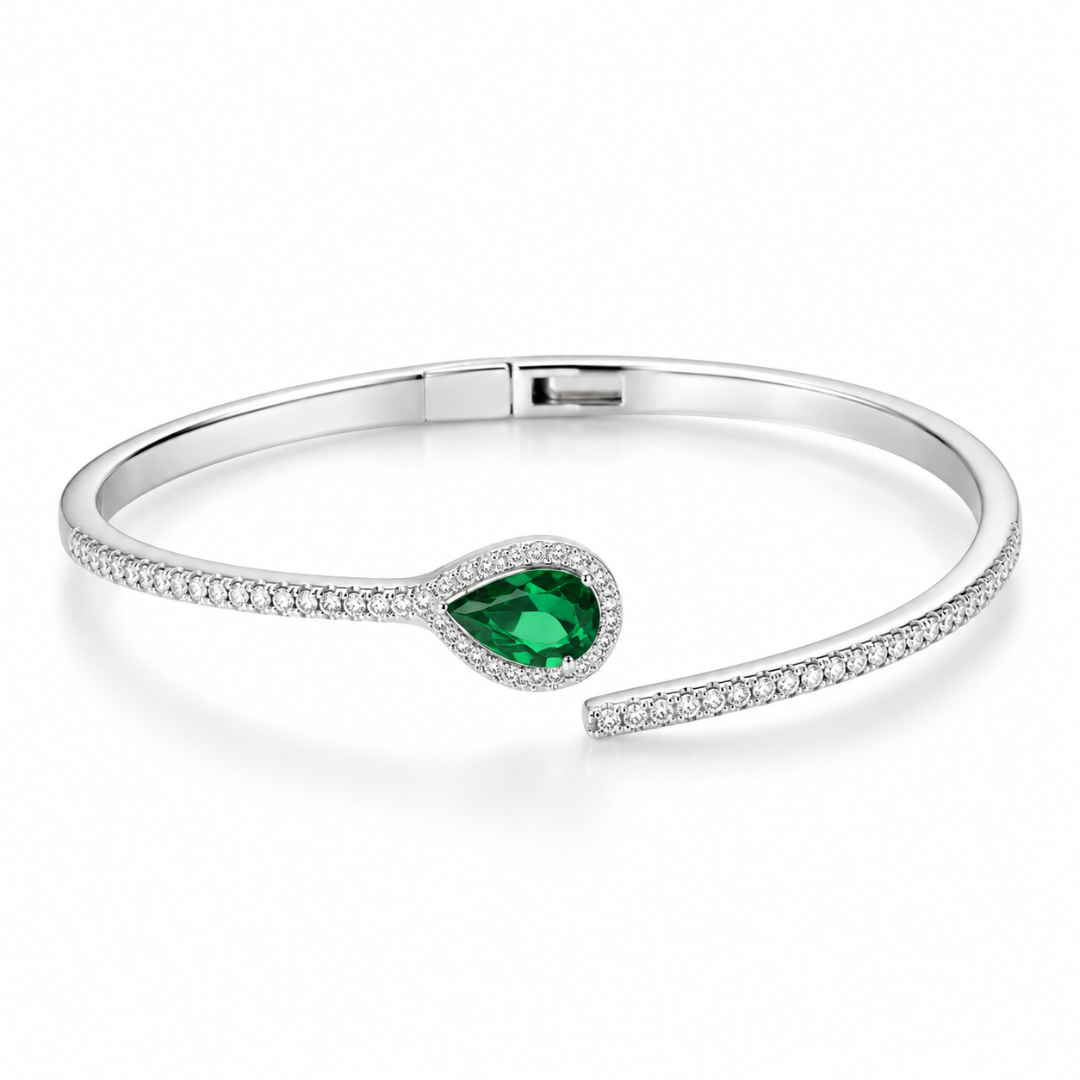 A polished silver-toned open cuff bracelet with a teardrop-shaped green gemstone surrounded by small clear stones, displayed on a soft cream fabric background with a textured block and text reading 'FITS UP TO WRIST 2.8'.