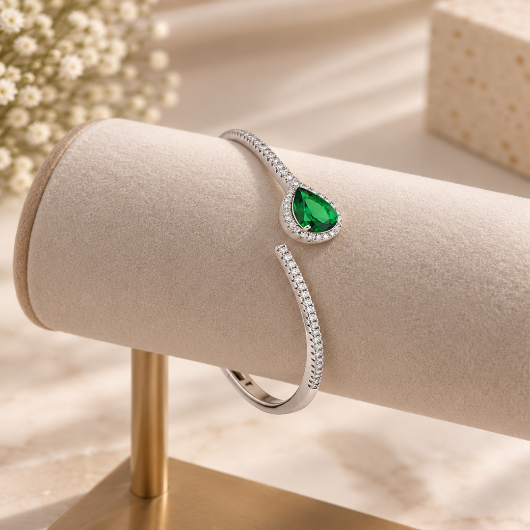 A silver-toned open cuff bracelet with a teardrop-shaped green gemstone and small clear stones, displayed on a beige jewelry stand.