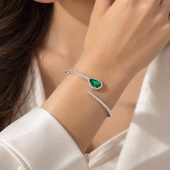 A close-up of a woman's wrist wearing a silver cuff bracelet with a teardrop-shaped green gemstone and a halo of small clear stones, paired with a matching slim silver band.