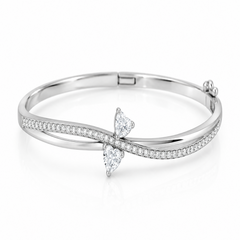 A silver-toned hinged bangle bracelet featuring a bow-shaped setting with two clear heart-cut gemstones and a row of small pavé-set stones along the curved upper band, shown on a white background.