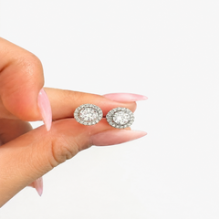 A close-up of a hand with pink manicured nails holding a pair of round halo stud earrings with clear gemstones on a plain white background.