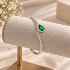 A silver-toned open cuff bracelet with a teardrop-shaped green gemstone and small clear stones, displayed on a beige jewelry stand.